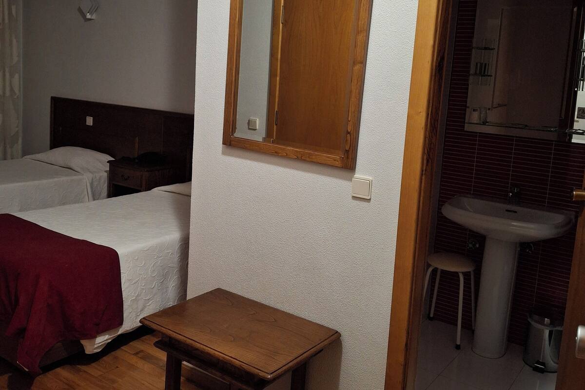 A hotel room with two single beds, a small wooden table, a mirror on the wall, and a bathroom visible through an open door.