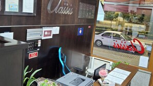Un mostrador de recepción con una computadora, una impresora y una planta. Se ve un coche y un edificio a través de la ventana.