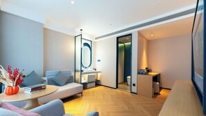 Une chambre d’hôtel moderne avec un canapé, une petite table surmontée d’un vase, un miroir, un plancher en bois et une vue sur la salle de bain.