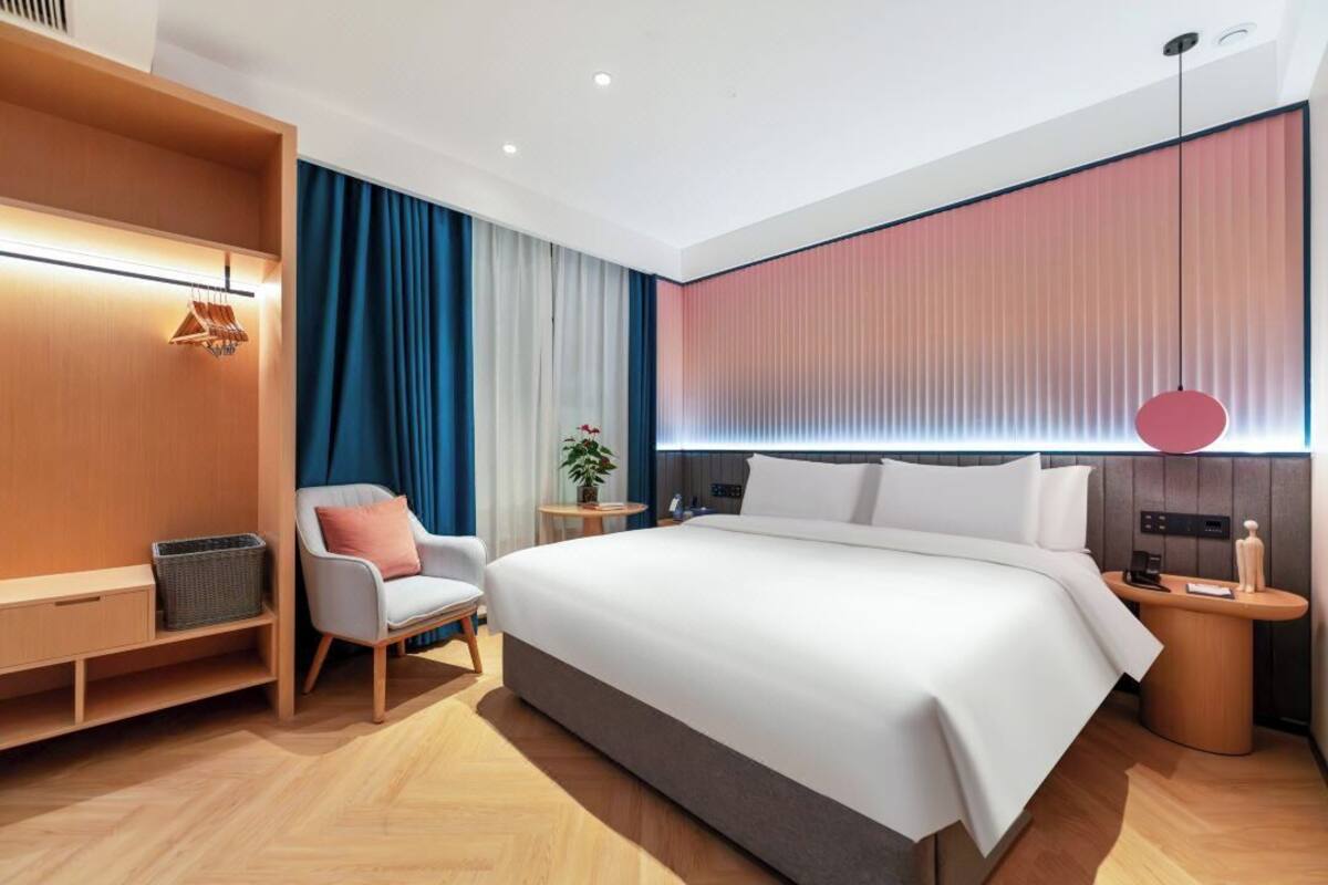 Une chambre d’hôtel moderne avec un grand lit, un coin salon comprenant un fauteuil, une petite table et une lampe fixée au mur.