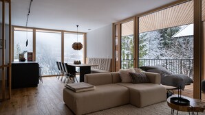 A modern living room with a large sectional sofa, a dining area with a table and chairs, and large windows offering a view of snow-covered trees.