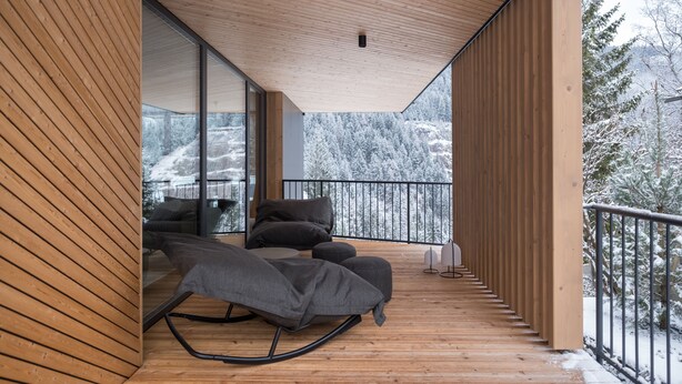 A wooden deck with a sofa and pillows, large windows, and a view of snow-covered trees.