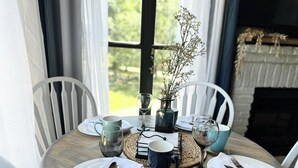 Mesa de jantar posta para quatro, com pratos brancos, talheres e copos azuis e verdes. Um vaso com flores secas está sobre a mesa, e visível ao fundo, uma lareira.