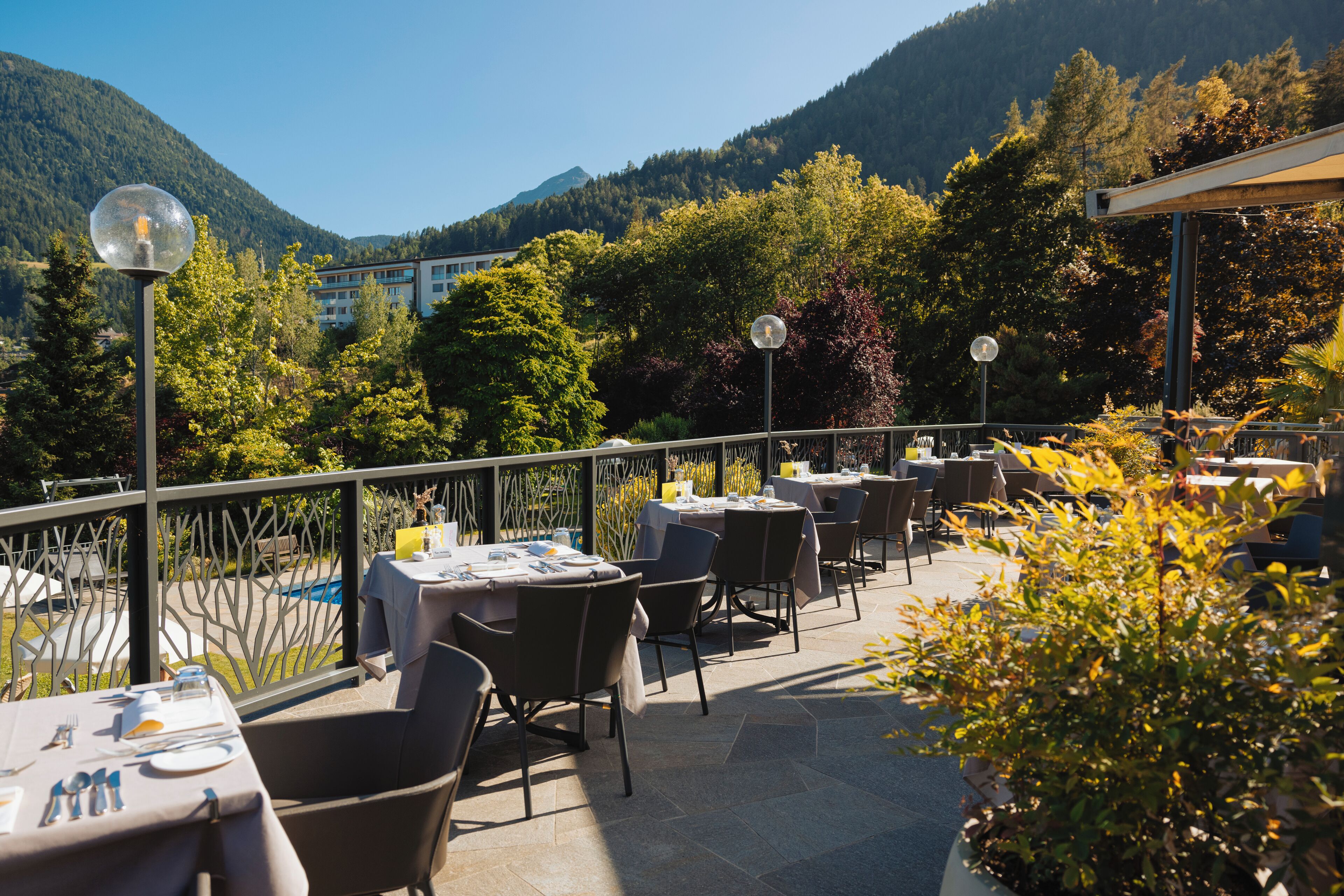 Un área de comedor al aire libre con mesas y sillas, rodeada por una barandilla metálica, con vista a una piscina y colinas cubiertas de bosque.
