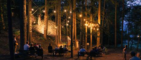 A group of people seated on benches in a forested area with string lights.