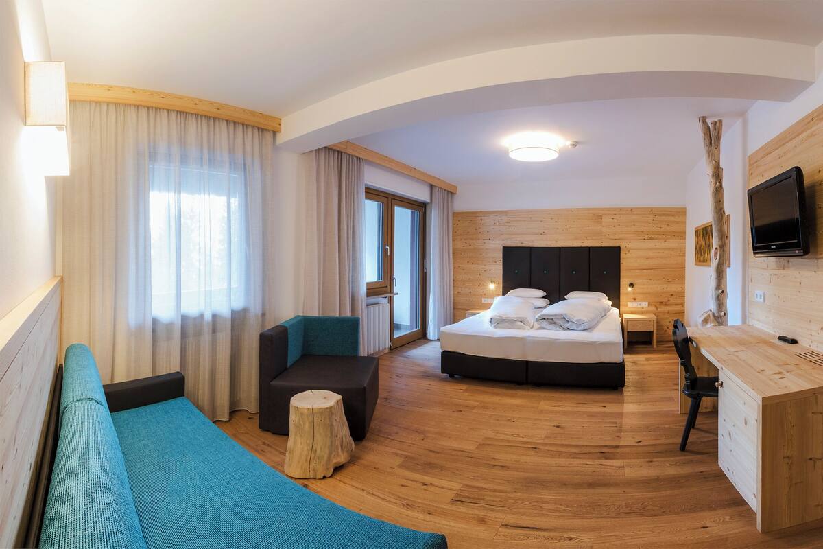 A modern hotel room with a wooden floor, a bed with white linens, a desk, a TV mounted on the wall, and a large window with curtains.
