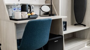 Kompakti hotellihuone, jossa on työpöytä, tuoli, televisio, vedenkeitin, kahvinkeitin, vesiautomaatti ja pieni musta roskakori.