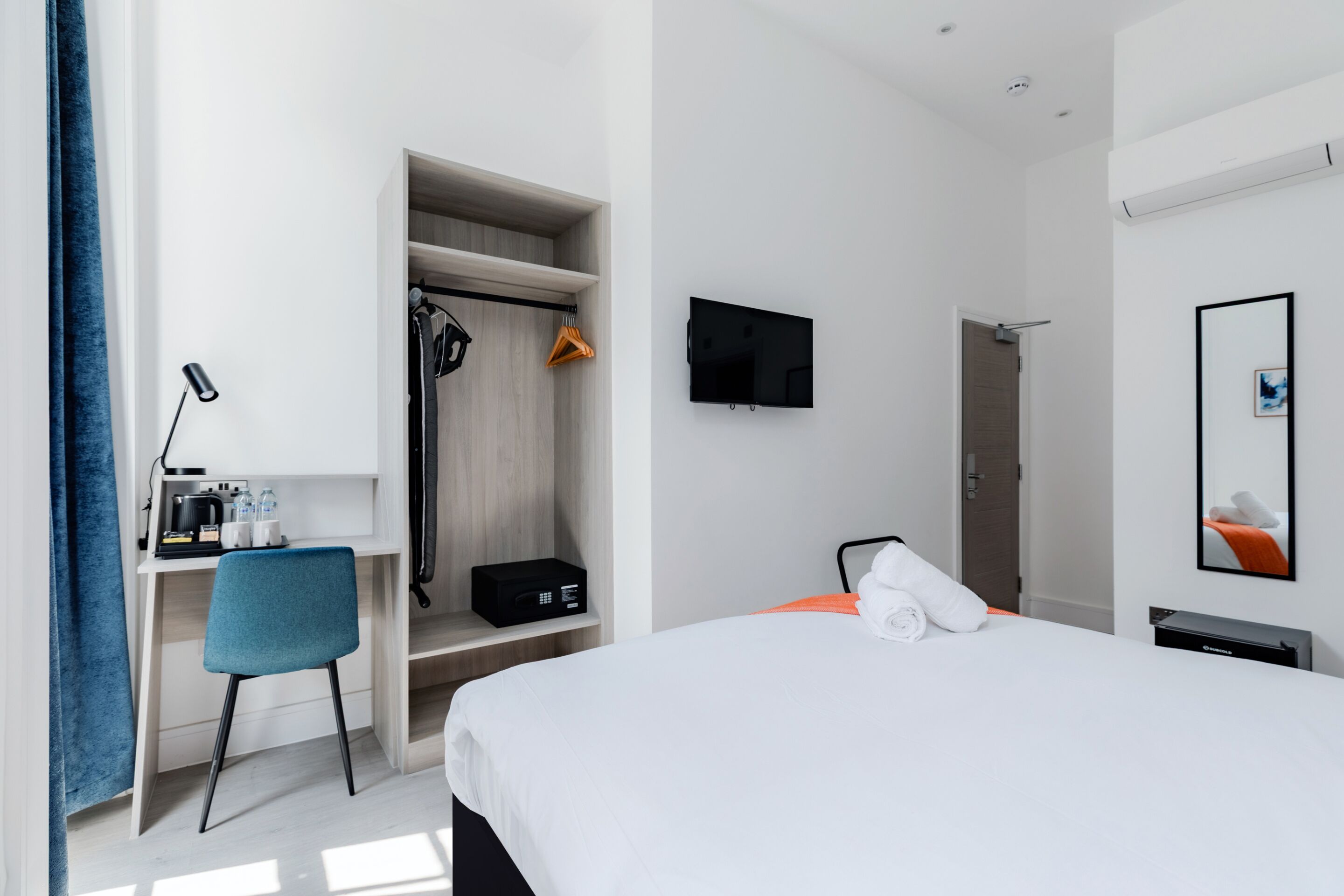 A hotel room with a bed, a desk with a chair, a TV, a wardrobe, and a shower area.