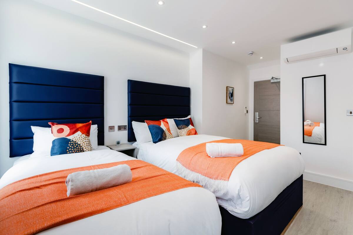 A hotel room with two beds, each with a blue headboard, white bedding, and orange accents.