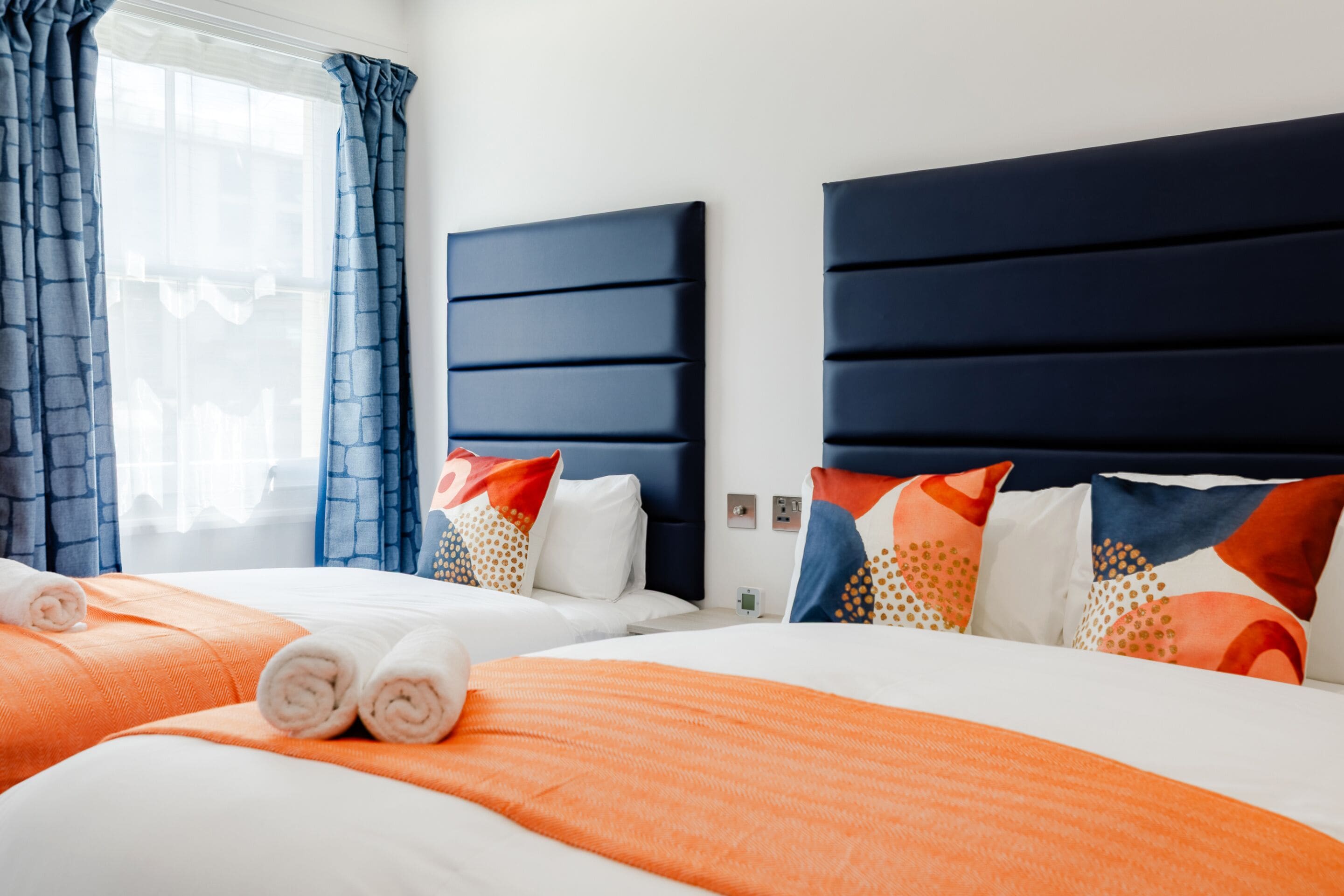 A hotel room with two beds, each with a blue headboard, white bedding, and orange accents.