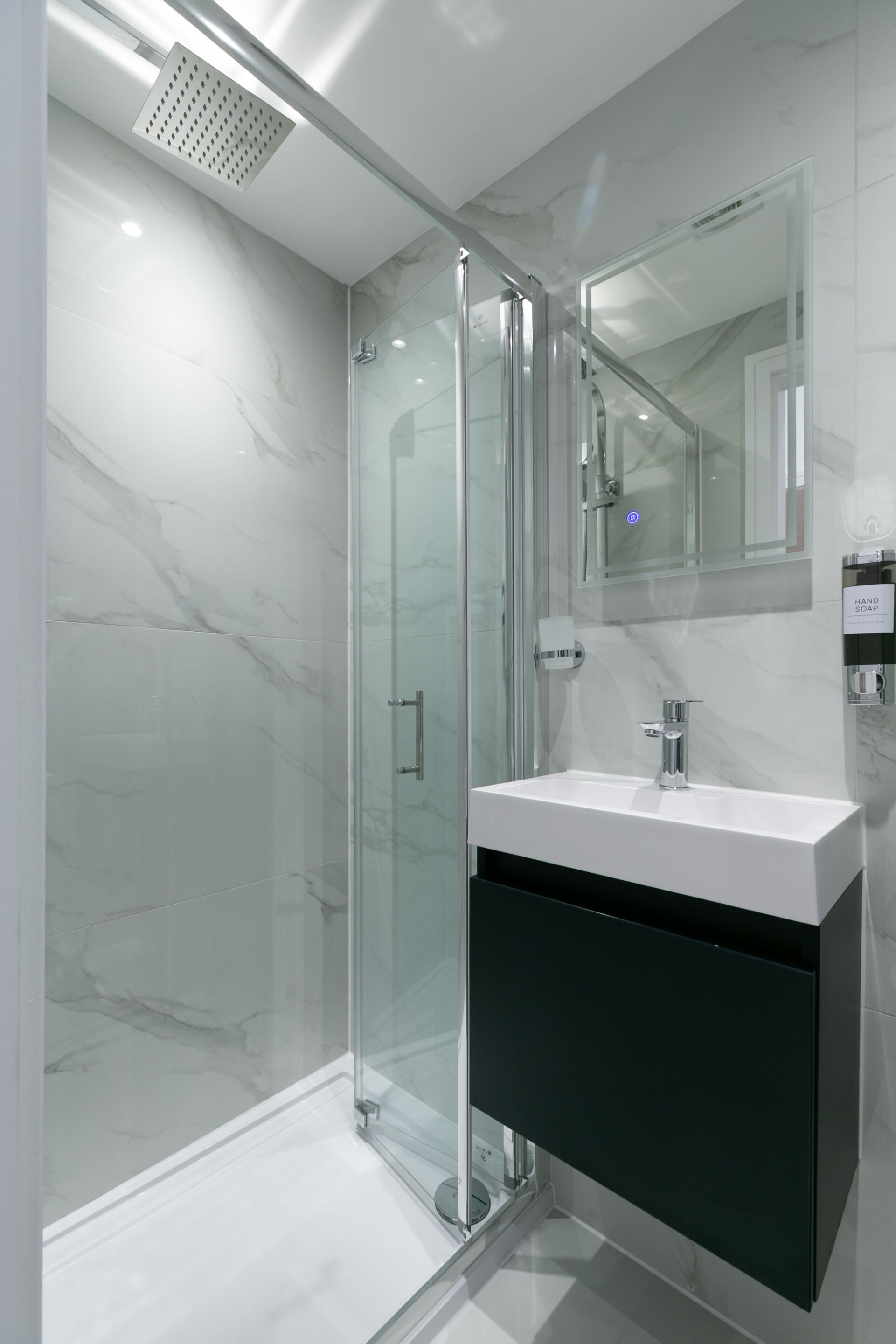 Une salle de bain moderne avec une douche à l’italienne en verre, un lavabo blanc avec un meuble noir et un distributeur de savon mural.