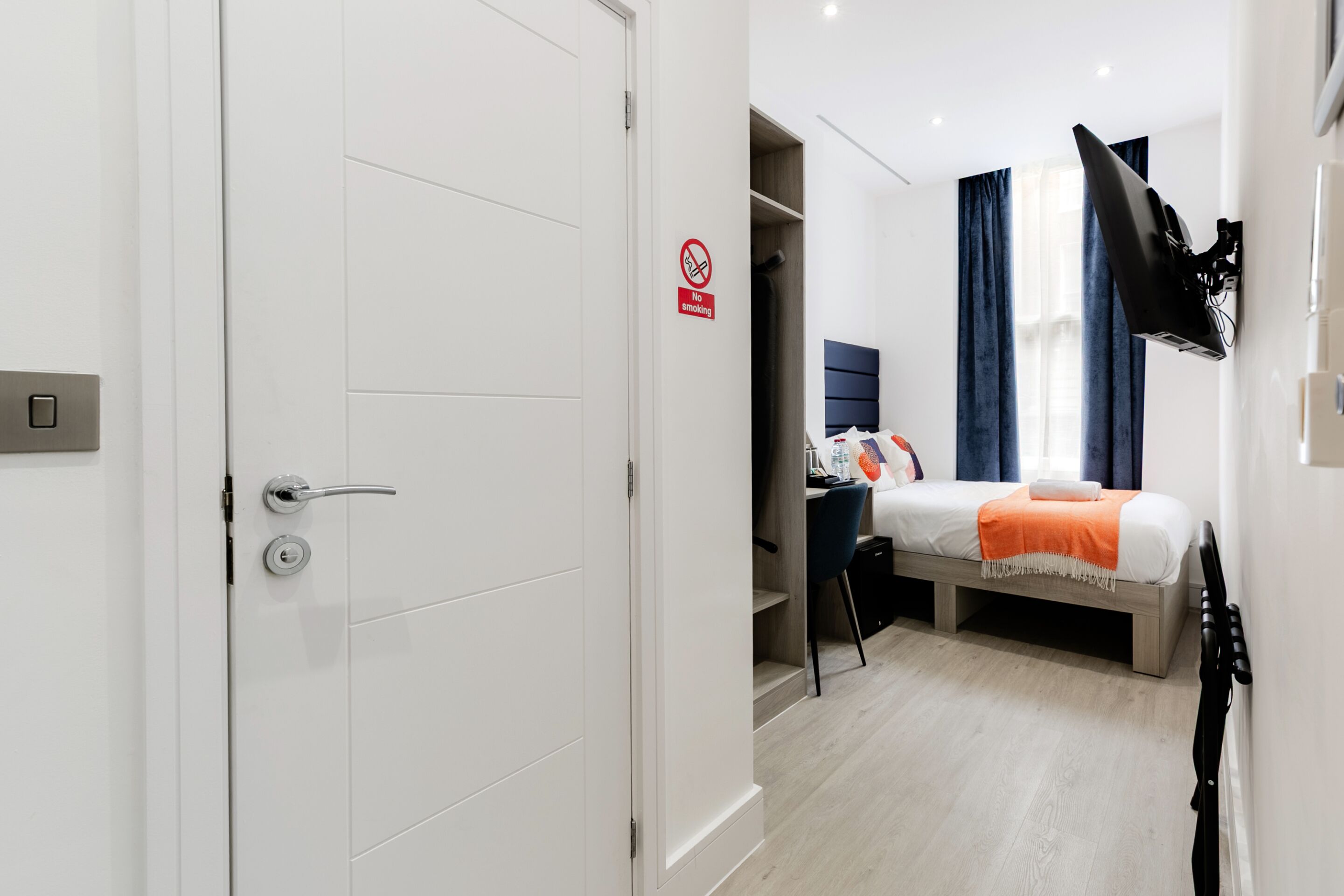 A hotel room with a bed, a desk, a chair, a TV mounted on the wall, and a no smoking sign.