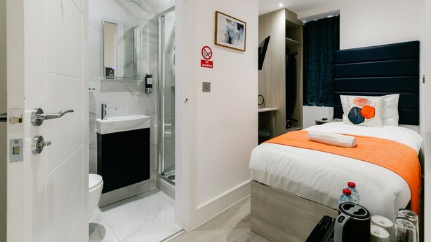 A modern hotel room with a bathroom, a bed with an orange bedspread, a no smoking sign, and a framed picture on the wall.