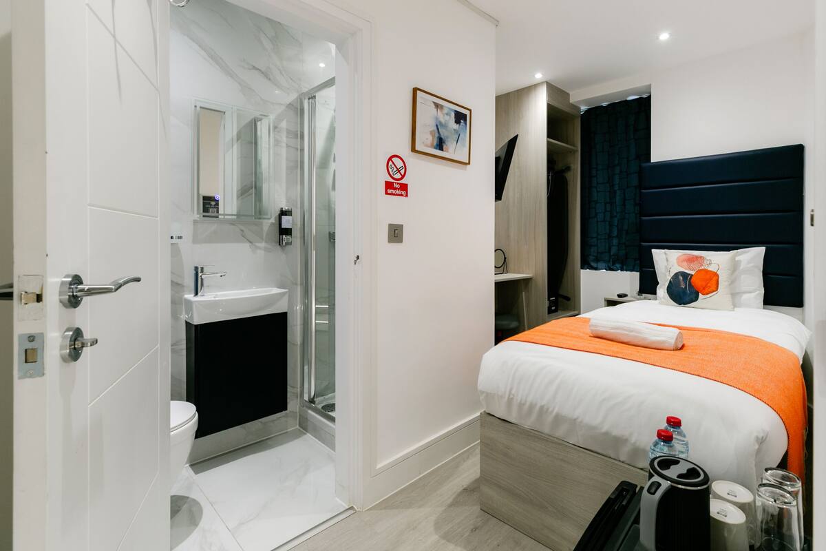 A modern hotel room with a bathroom, a bed with an orange bedspread, a no smoking sign, and a framed picture on the wall.