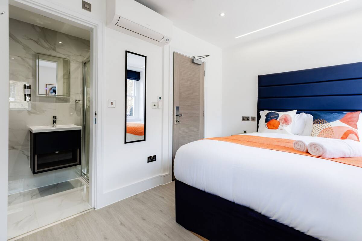 A modern bedroom with a large bed, a bedside table, a mirror, and a bathroom with a sink and shower.