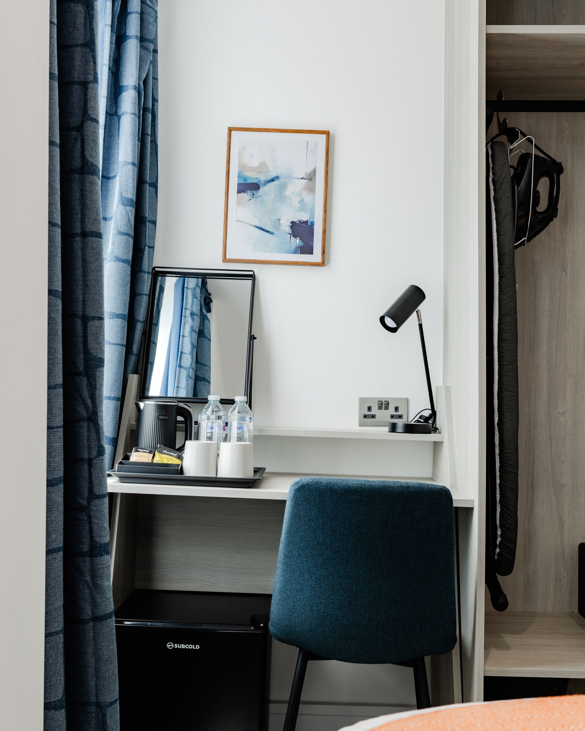 A hotel room with a desk, a mirror, a coffee maker, a water bottle, a framed abstract painting, and a blue chair.