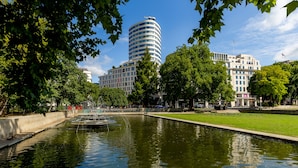Puisto, jossa on suihkulähde, lampi ja taustalla moderneja rakennuksia.