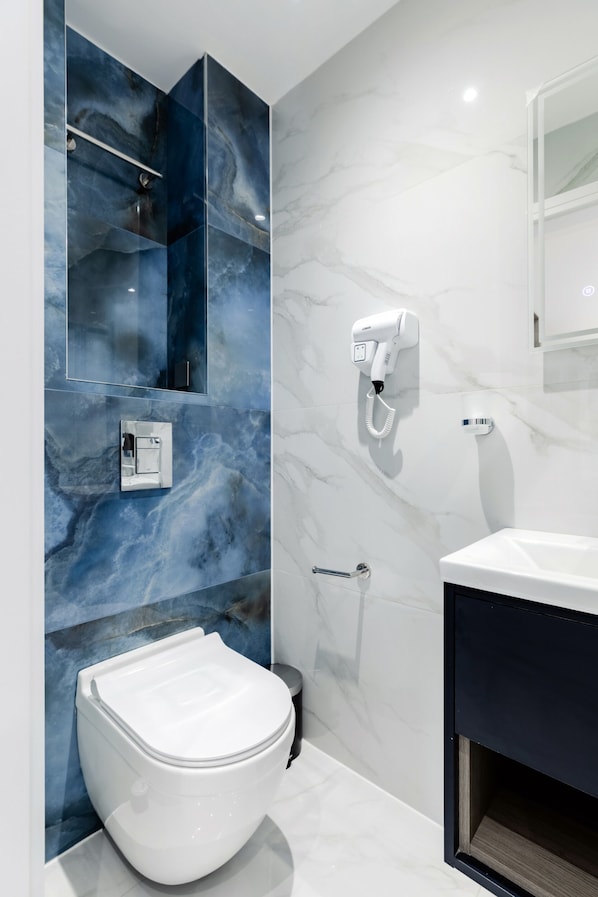 A modern bathroom with a marble wall, a white toilet, a built-in sink, and a hairdryer mounted on the wall. - Upper Berkeley Hotel (London)