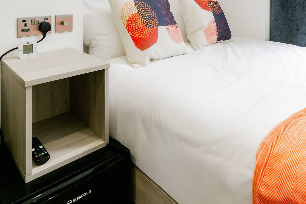 A hotel room with a bed, a bedside table with a remote control, a nightstand, a wall socket, and a power outlet.
