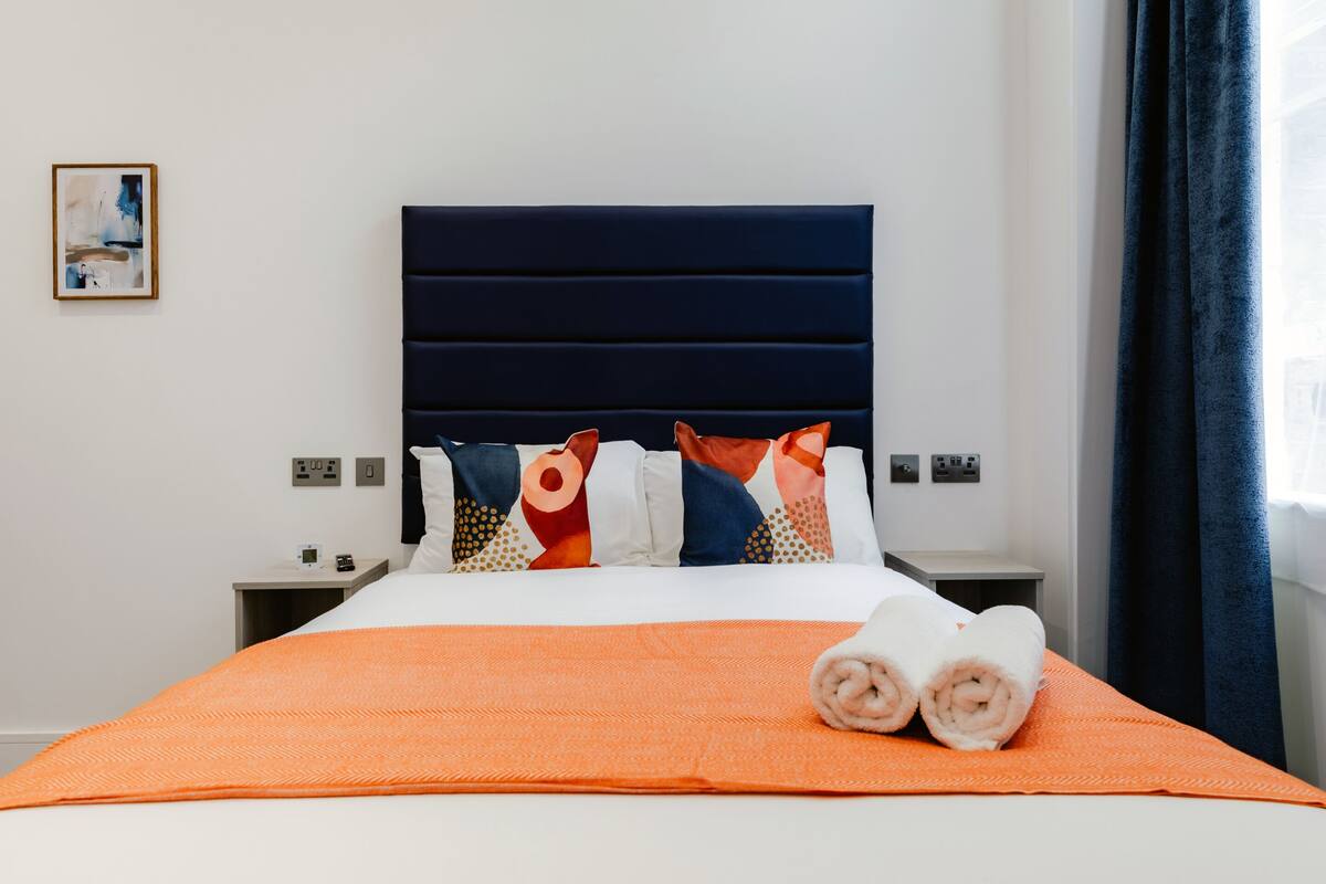 A hotel room with a large bed, an orange blanket, two rolled towels, a dark blue headboard, and a painting on the wall.