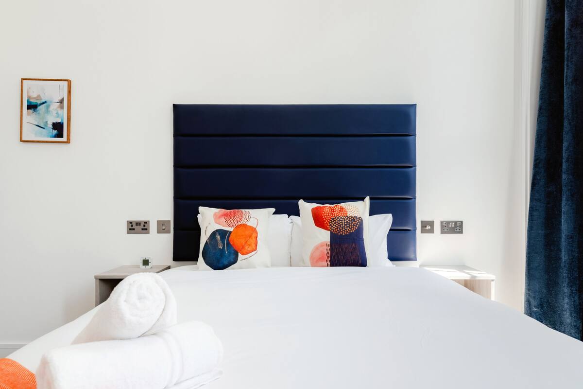 A modern hotel room with a large bed, a dark headboard, and a framed abstract painting on the wall.