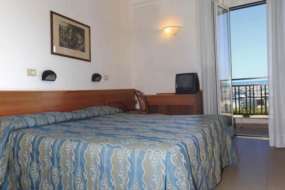 A hotel room with a bed, a desk, a chair, a television, and a balcony with curtains.