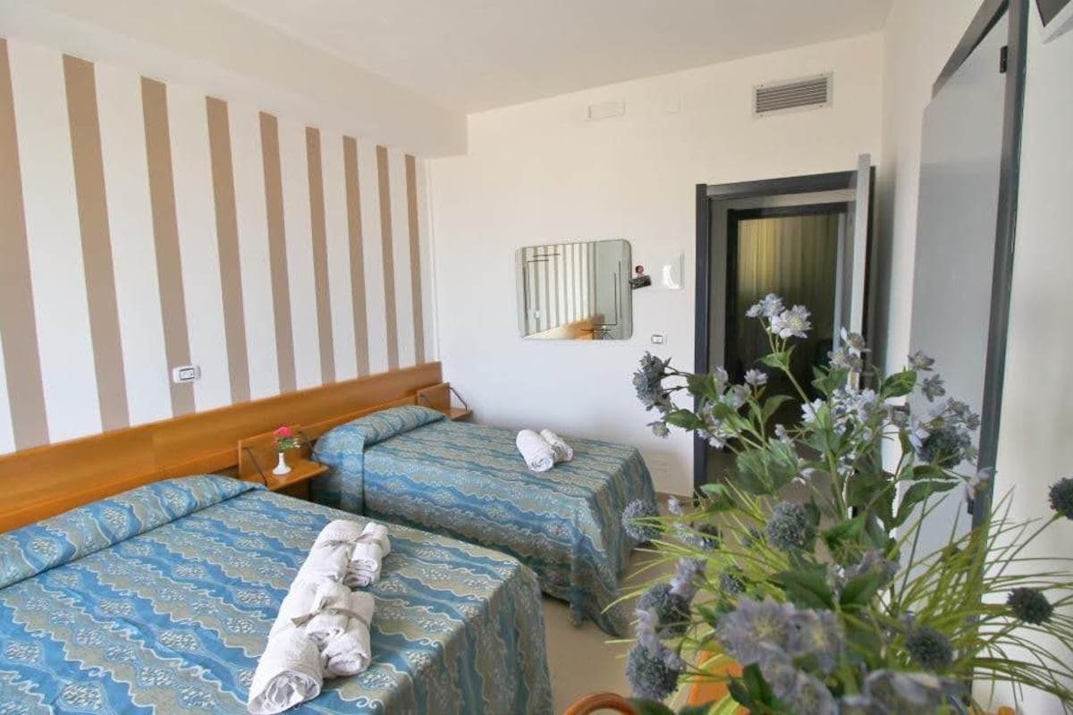 A hotel room with two beds, a wooden headboard, a small bedside table with a vase, a mirror, and a potted plant.