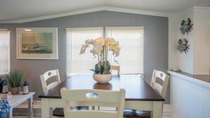 A dining area with a dark wooden table, white chairs, and a vase of white orchids. There are framed pictures on the wall and a window with blinds. - 2 BR@Peachtree, Walk2Sand + Sit by Water (Ocean City)