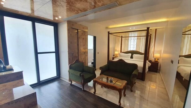A modern hotel room with a large bed, a sitting area with two armchairs, a glass coffee table, and a large window providing natural light. - Hotel Daffodils Inn (Dehradun)