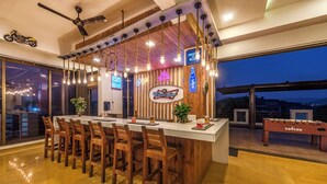 Un espace bar avec des tabourets de bar en bois, un comptoir de bar blanc, des luminaires suspendus, des enseignes au néon et une table de baby-foot.