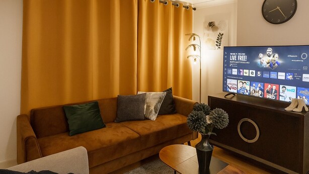 Een woonkamer met een bruine bank, een bijzettafeltje, een televisie op een kast en een klok aan de muur.