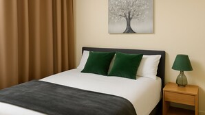 Een netjes opgemaakt bed met een donker hoofdbord, twee groene kussens en een wit kussen. Een groene lamp staat op een houten nachtkastje naast het bed.