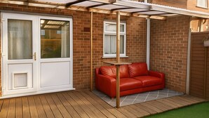 Een terras met een houten terrasvloer, een rode leren bank en een witte deur met glaspanelen.