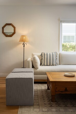 A living room with a sofa, a wooden coffee table, a floor lamp, a patterned ottoman, and a window with blinds. - Carlsbad Cottage - Remodeled (Carlsbad)