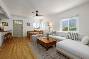 A spacious living room with a white sofa, wooden coffee table, and a dining area with a wooden table and chairs. - Carlsbad Cottage - Remodeled (Carlsbad)