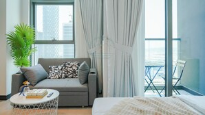 A modern hotel room with a grey sofa, a small round coffee table, a bed, and a balcony with a view of the city.