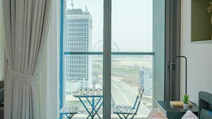 A hotel room with a large window, a view of a cityscape, a small table with chairs, a bedside lamp, and a potted plant.