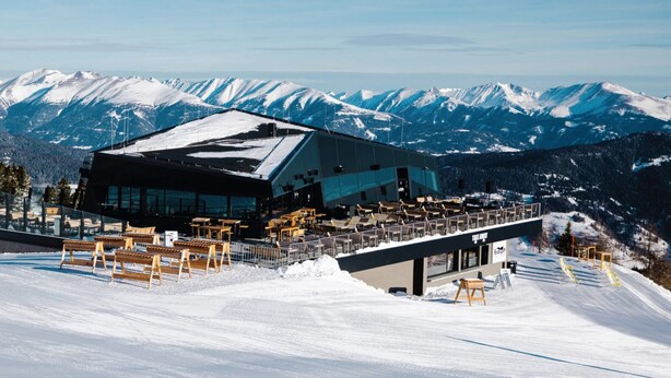 A modern ski resort building with a glass facade, outdoor seating area, and a view of snow-covered mountains. - Apartment in Kreischberg Near Ski Resort (Sankt Georgen am Kreischberg)