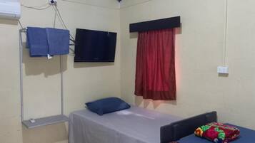 A small, single-bed room with a TV mounted on the wall, a window with red curtains, and a small table with a colorful cushion.
