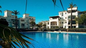 A swimming pool surrounded by multi-story buildings with balconies, palm trees, and a clear blue sky. - Stunning 2-baths apartment with golf and lake view (Sucina)