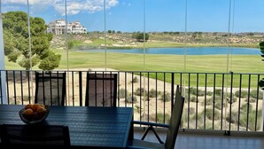 A view from a balcony with a table and chairs, overlooking a golf course and a body of water, with a building in the background. - Stunning 2-baths apartment with golf and lake view (Sucina)
