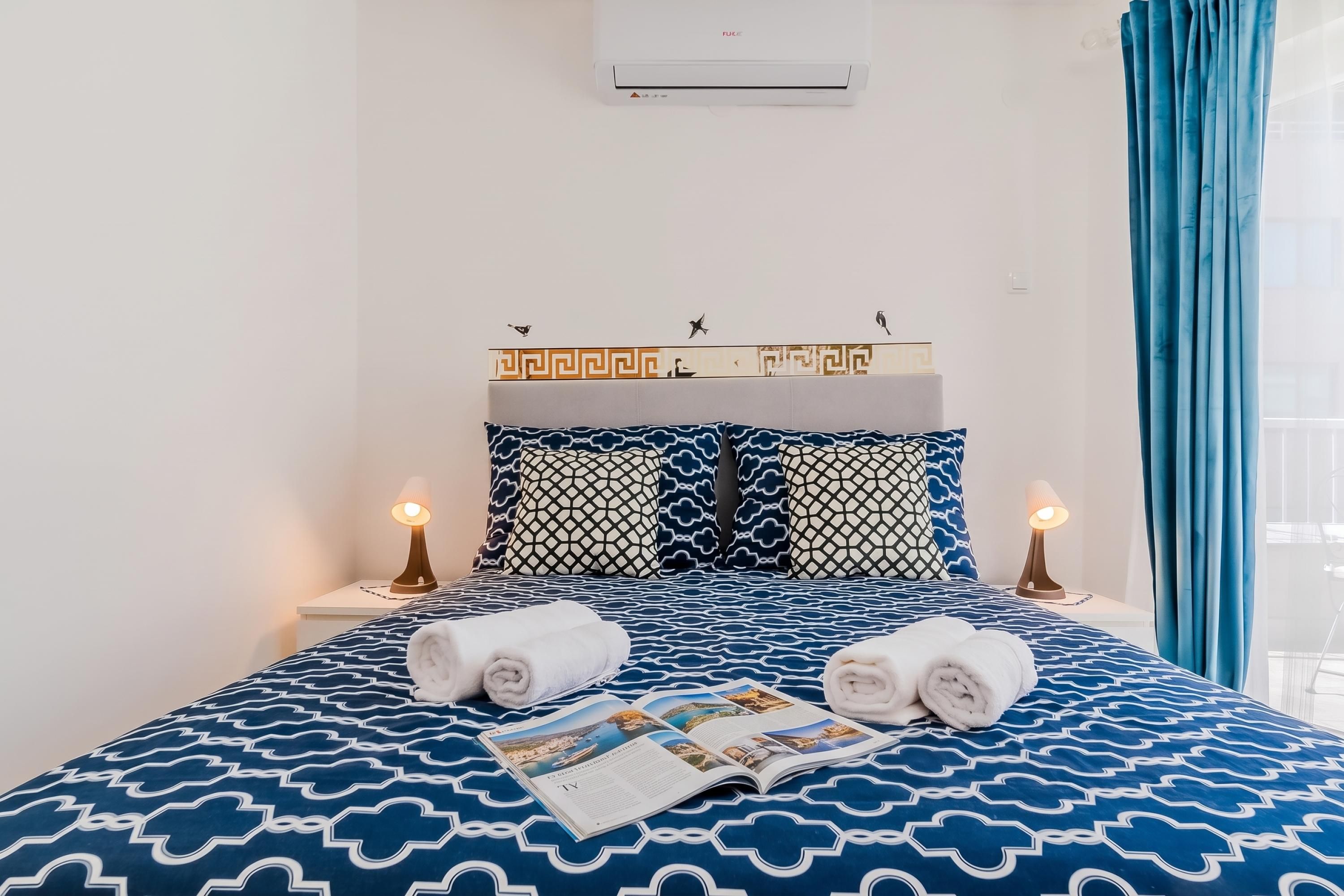 Een hotelkamer met een bed, twee bedlampen, een raam met blauwe gordijnen en een airconditioner aan de muur.