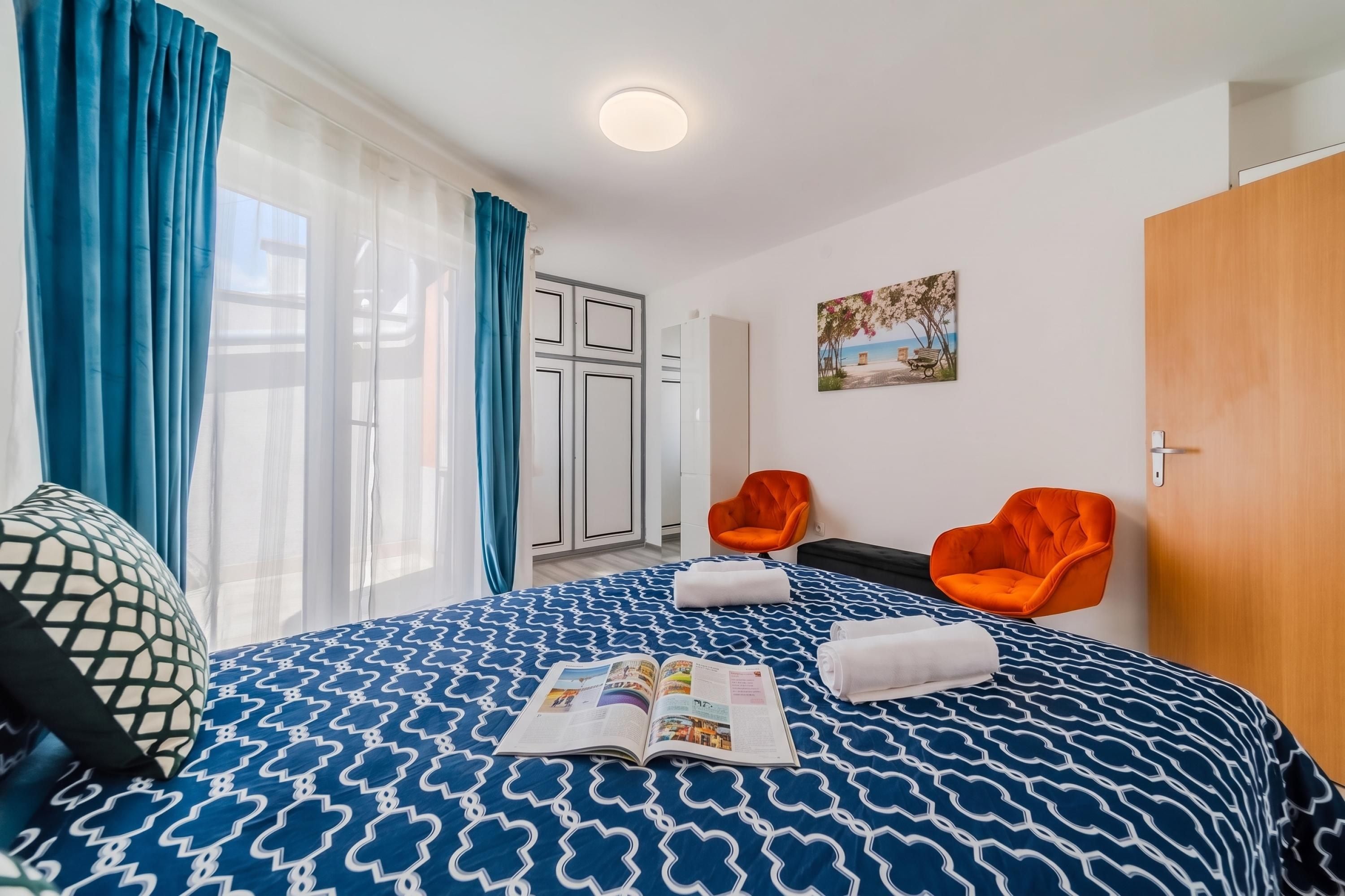 A hotel room with a bed, two orange chairs, a wooden door, and a painting on the wall.