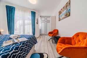 A hotel room with a bed, a desk, an orange chair, a black chair, a painting, and a door.