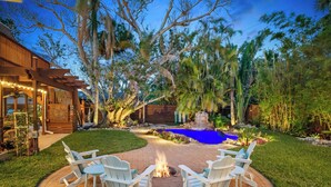 A patio with a fire pit and Adirondack chairs, surrounded by a pool and lush greenery. - Walk to Gulf: Luxe Luau-themed Gem in Holmes Beach (Holmes Beach)