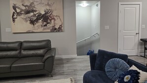 A living room with a gray leather couch, a blue armchair with decorative pillows, a large abstract painting on the wall, and a staircase leading to a second floor.