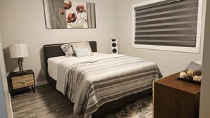 A bedroom with a bed, a bedside table with a lamp, a wall-mounted speaker, a framed picture, a window with blinds, and a decorative rug.
