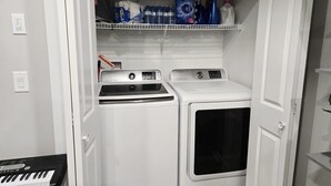 A compact laundry room with a washing machine and dryer, a shelf with cleaning supplies, and a keyboard on the floor.