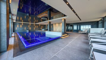 Ein moderner Poolbereich im Innenbereich mit einem blau beleuchteten Whirlpool, Liegestühlen und einer Rezeption.