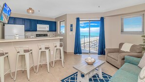 A modern living room with a balcony, a kitchen with blue cabinets, and a dining area with white chairs. - Myrtle Beach Resort RT1112 (Myrtle Beach)